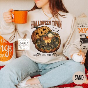 Pode incluir: Uma camisola de gola redonda cor de areia com um gráfico de uma grande abóbora esculpida com uma cena urbana no interior. O texto diz HALLOWEEN TOWN e EST. 1998. O design inclui silhuetas de figuras, uma lua cheia e morcegos ao pôr do sol.