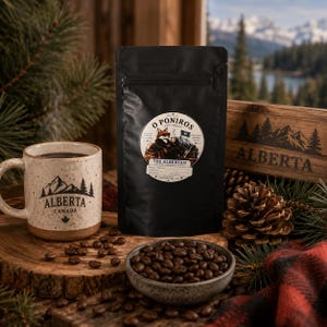Könnte beinhalten: Eine schwarze Kaffeetüte mit einem runden Etikett, das einen Fuchs und den Text O Poniros The Albertan zeigt. Daneben stehen eine gesprenkelte Keramiktasse mit einem Alberta Canada Bergmotiv und eine Schale mit gerösteten Kaffeebohnen.