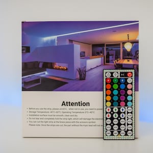 May include: A rectangular product box for LED light strips featuring a room interior image and an Attention section with usage instructions. A black remote control with forty-four colorful buttons is positioned in front of the box.