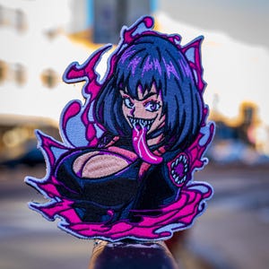 May include: An embroidered patch featuring a stylized character with dark hair, sharp teeth, and a long pink tongue. The character is surrounded by vibrant pink liquid-like shapes and accents, set against a dark background with a white border.