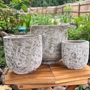 May include: A set of three cylindrical planters with a rough, weathered, stone-like texture in shades of gray and brown. The planters vary in height and width, featuring a wide opening at the top and a slightly tapered base, arranged together.