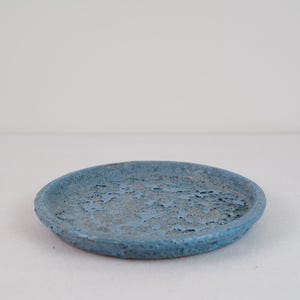 May include: A circular ceramic tray featuring a textured, porous surface with a mottled blue and grey finish. The edges are slightly raised, and the overall appearance is rugged with a matte, uneven glaze that highlights the natural material.