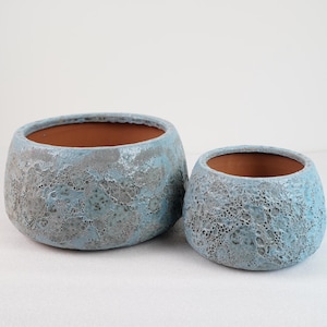 May include: Two round ceramic planters with a textured, cratered surface in shades of light blue and grey. The exterior features a porous, uneven finish, while the interior of each bowl-shaped vessel reveals a smooth, terracotta-colored clay.