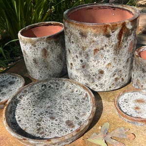 May include: A collection of three cylindrical ceramic planters and two matching circular saucers. Each piece features a mottled, speckled glaze in shades of white, gray, and brown, with a smooth terracotta-colored interior and rim.