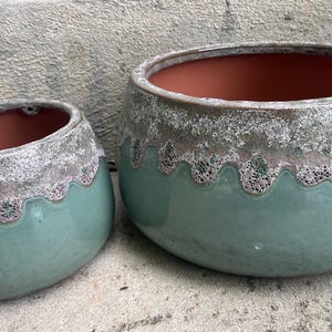 May include: Two round ceramic planters with a smooth, glossy sage green base and a textured, mottled white and brown glaze along the upper rim. The interior of both pots is a solid terracotta orange color, featuring a distinct scalloped edge design.