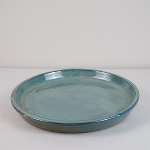 May include: A round ceramic plate with a glossy finish, featuring a gradient of blue and teal tones that transition into subtle earthy brown hues along the edges and the base. The surface is smooth with a slightly raised rim.