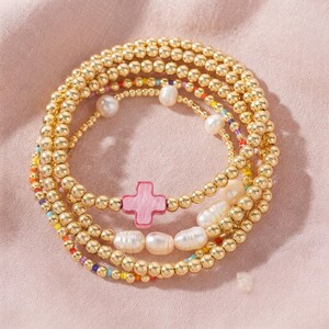 May include: A stack of four bracelets featuring polished gold-tone metallic beads, white freshwater pearls, and a central pink marbled cross charm. The design incorporates small, multi-colored seed beads interspersed throughout the metallic strands.