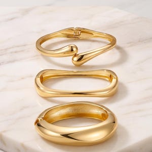 May include: Three gold-toned metal hinged bracelets with polished finishes. The top bracelet features a bypass design with teardrop ends, the middle is an elongated oval shape, and the bottom is a thick, rounded band with a smooth, reflective surface.