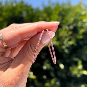 May include: A pair of thin hoop earrings featuring a row of small, vibrant pink gemstones set in a metallic frame. The earrings are circular in shape and display a consistent pattern of pink stones along the entire visible length of the thin metal band.