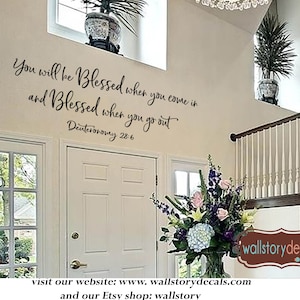 May include: A white wall with a black vinyl decal that reads "You will be Blessed when you come in and Blessed when you go out Deuteronomy 28:6". The decal is placed above a white door with a glass window. A wooden staircase with a white railing is to the right of the door. A bouquet of flowers is on the bottom step of the staircase.
