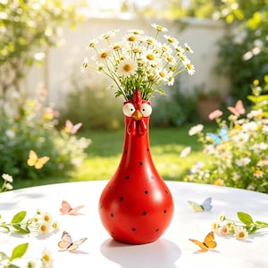 Puede incluir: Un jarrón de cerámica rojo brillante con forma de gallina, lunares negros, ojos blancos grandes y pico amarillo. El jarrón contiene un ramo de margaritas blancas con centros amarillos, sobre una superficie blanca con mariposas dispersas.