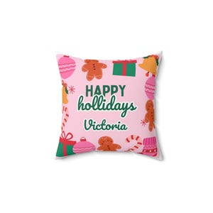 May include: A square throw pillow with a light pink background featuring a repeating pattern of gingerbread people, green and red gift boxes, pink ornaments, candy canes, and bells. The text "HAPPY hollidays" and "Victoria" is in dark green.