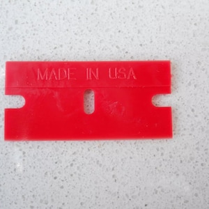 May include: A red plastic scraper with a handle and a blade. The scraper has the words "MADE IN USA" printed on it.