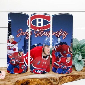 Könnte beinhalten: Ein zylindrischer Becher mit einem Grafikdruck eines Eishockeyspielers im rot-blauen Trikot, dem Logo der Montreal Canadiens und dem Namen Juraj Slafkovsky in roter Schreibschrift vor einer dunkelblauen nächtlichen Skyline.