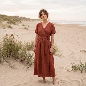 May include: A rust-colored midi-length wrap dress featuring short sleeves, a V-neckline, and a matching fabric tie belt at the waist. The garment is constructed with a simple, clean design, showcasing a solid earthy tone and a relaxed silhouette.