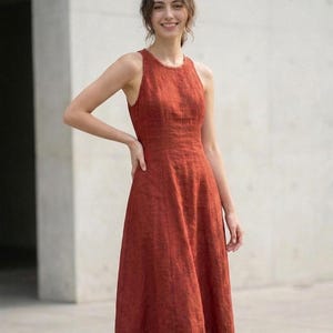 May include: A sleeveless midi dress in a deep rust-orange color. The garment features a high, rounded neckline with a subtle textured finish, a fitted bodice with vertical seam detailing, and a flared skirt that falls below the knee.