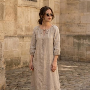 May include: A beige midi-length dress with three-quarter length sleeves, a round neckline, and a front tie closure. The garment features a loose, relaxed fit with gathered detailing at the neckline and cuffs, crafted from a textured fabric.