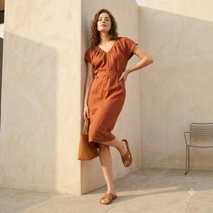 May include: A knee-length, rust-colored dress with short sleeves, a V-neckline, and a defined waistline. The garment is paired with brown leather slide sandals and a matching brown leather tote bag, creating a cohesive look with simple lines.