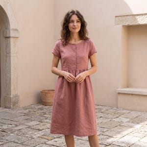 May include: A dusty rose midi dress featuring short sleeves, a round neckline, and a front button placket extending to the waist. The garment has a fitted bodice with a gathered skirt that falls to the mid-calf, crafted from a solid-colored fabric.