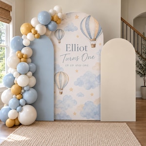 May include: A party backdrop set featuring three arched panels, one with a hot air balloon and cloud print reading "Elliot Turns One UP UP AND ONE," accompanied by a large balloon garland in shades of blue, white, and mustard yellow.