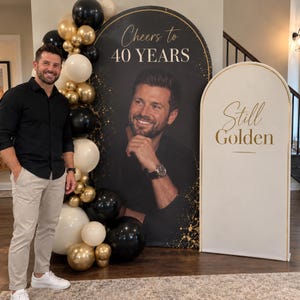 Puede incluir: Un gran telón de fondo arqueado negro con el texto Cheers to 40 YEARS y el retrato de un hombre, acompañado de un arco blanco más pequeño con el texto Still Golden, decorado con un racimo de globos negros, dorados y de color crema.