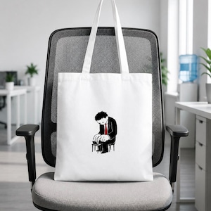 Può includere: Una borsa tote in tela bianca con un'illustrazione grafica centrata di una persona in abito nero e cravatta rossa seduta su un piccolo sgabello mentre digita su un computer portatile. La borsa è dotata di due lunghi manici bianchi.
