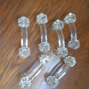 May include: A set of six clear glass drawer pulls, each featuring a curved handle design with hexagonal, faceted ends. The pulls have a transparent, reflective surface and a central mounting hole on each of the two hexagonal end pieces.