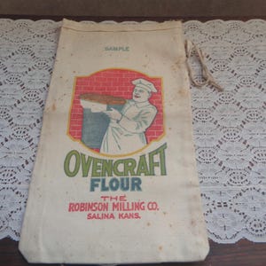 May include: Vintage flour sack with a drawstring closure. The sack features a colorful graphic of a chef holding a tray of baked goods, with the words "OvenCraft Flour" and "The Robinson Milling Co." printed on the front.