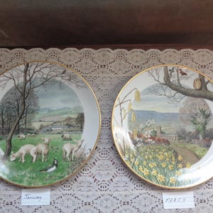 May include: Two decorative plates with gold rims. One plate depicts sheep in a green field with a tree, labeled "January." The other plate shows a path with yellow flowers and a church, labeled "March."