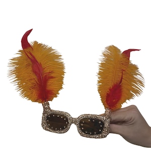 May include: Novelty glasses featuring a gold-toned frame covered in small sparkling crystals. Each lens frame is topped with a large, upright plume of bright orange feathers accented with vibrant red feathers extending from the center.