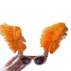 May include: Sunglasses featuring a frame covered in small, light-colored rhinestones. Two large, vibrant orange feathers extend upward from the top corners of the frames. The lenses are dark, and the overall design is symmetrical and decorative.