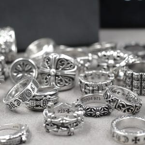 May include: A collection of silver rings featuring intricate embossed patterns, including floral motifs, cross designs, and engraved text such as 1994 925 and Forever. The rings are crafted from polished sterling silver with dark oxidised detailing.