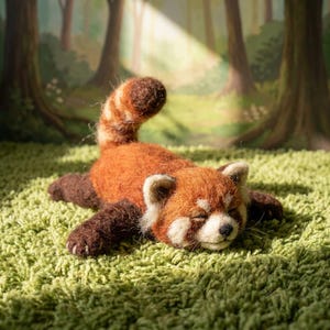 May include: A needle-felted red panda figurine with a rust-orange back, dark brown limbs, and a striped tail. The creature has white facial markings, a black nose, and closed eyes, depicted in a resting position with its body stretched out.