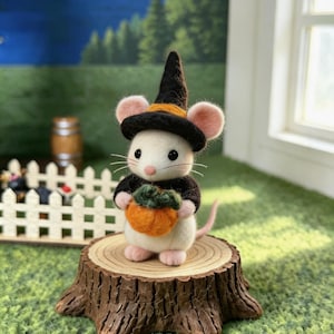 Puede incluir: Una figura de ratón blanco de lana fieltrada que lleva un sombrero de bruja negro con una banda naranja y un suéter negro. El ratón sostiene una pequeña calabaza naranja con un tallo verde y se apoya sobre una base de tronco de árbol.