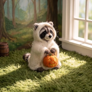 Puede incluir: Una figura de mapache de lana fieltrada con una aguja que lleva un disfraz de fantasma blanco. El mapache tiene pelaje oscuro alrededor de los ojos y sostiene una pequeña calabaza de Halloween naranja de fieltro con una cara tallada.