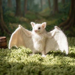 May include: A needle-felted white bat figurine with outstretched wings, textured fur, and small dark eyes. The creature stands upright with its wings fully extended, showcasing the detailed fiber construction and soft, off-white color palette.