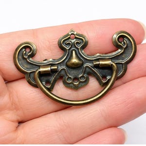 May include: An antique brass drawer pull with an ornate design. The pull features a central face-like motif, scrolled edges, and a curved handle. The hardware is held in a hand, showcasing its size and detail.