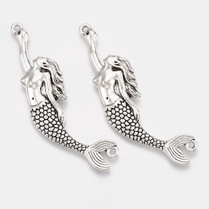 May include: Two silver mermaid charms with a raised design. The mermaids are facing to the right and have their arms raised above their heads. The charms have a loop at the top for attaching to jewelry.