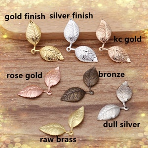 May include: Assortment of double leaf charms in various finishes, including gold, silver, rose gold, bronze, raw brass, and dull silver. The charms are displayed on a wooden surface.