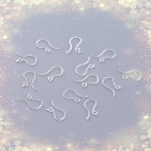 May include: A close-up of a group of clear plastic earring hooks. The hooks are arranged in a circle on a light gray surface.