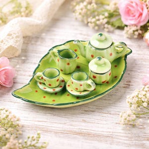 May include: A miniature ceramic tea set. The set includes a teapot, creamer, sugar bowl, and two cups and saucers. The set is light green with red polka dots and sits on a leaf-shaped tray. The background includes flowers and a white wooden surface.