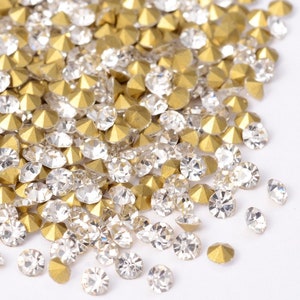 May include: A pile of clear and gold rhinestones, scattered on a white surface. The rhinestones are small and round, with a flat back and a pointed top.