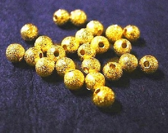 May include: A collection of small, round, gold-colored beads with a textured surface and a hole through the center. The beads are scattered on a dark blue surface, creating a contrast in color and texture. These beads are suitable for jewelry making.