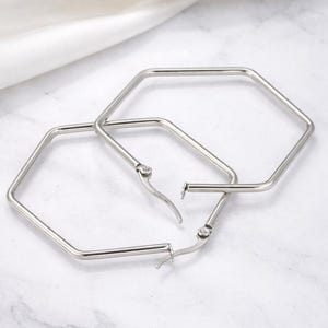 Op de afbeelding: Een paar zilverkleurige, zeshoekige oorringen. De oorbellen zijn gemaakt van glanzend metaal en hebben een eenvoudig, modern ontwerp. De oorbellen hebben een sluiting met een sluiting.