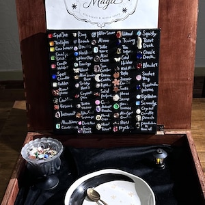May include: A wooden box containing a black display board with the text Joyside Magic and a list of items next to small charms. Below is a glass pedestal bowl filled with small trinkets, a white bowl with a black rim, and a metal spoon.