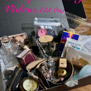 Op de afbeelding: Een zwarte kartonnen doos met een verzameling items, waaronder een doorzichtige glazen fles, een Modern Witch tarotdeck, een kleine metalen kelk, een kaars in een pot, een zwart etui met rits, een zilverkleurige ketting en diverse kleine snuisterijen.
