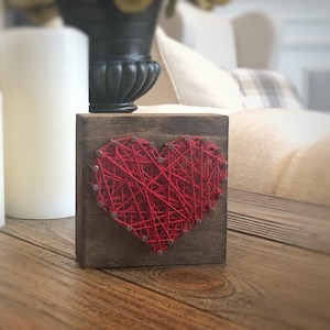 May include: A red heart-shaped string art piece on a small wooden block. The heart is created by wrapping red string around nails hammered into the wood.