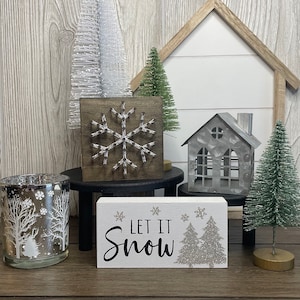 May include: A collection of winter-themed decorations, including a wooden sign that reads "Let It Snow" with glittery trees, a snowflake art piece, and miniature Christmas trees. A silver candle holder with a winter scene is also present.