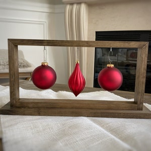 May include: Three red Christmas ornaments hanging from a wooden frame. The ornaments are a round ball, a teardrop shape, and another round ball. The frame is sitting on a white tablecloth.