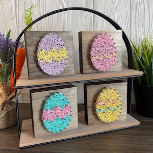 May include: Four wooden signs with string art of Easter eggs in pink, yellow, and blue. The signs are arranged in a tiered display on a black metal stand.